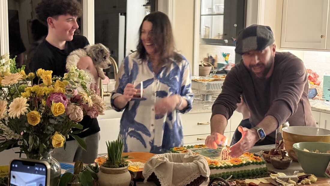 Anna Cunningham enjoying a celebration among family, friends and a dog in a kitchen with flowers, cake, chacuterie boards and other food items.
