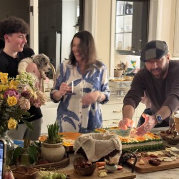 Anna Cunningham enjoying a celebration among family, friends and a dog in a kitchen with flowers, cake, chacuterie boards and other food items.