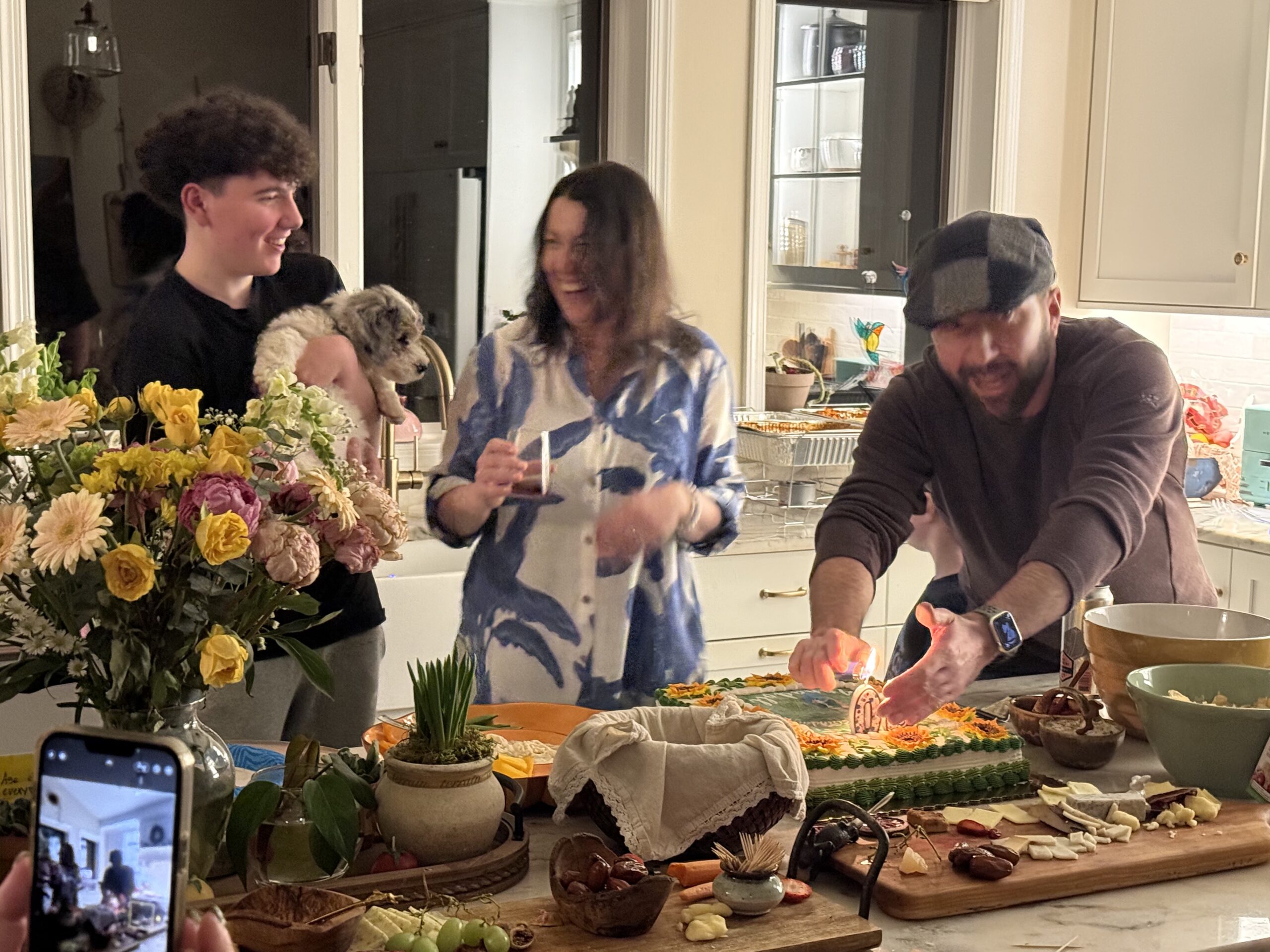 Anna Cunningham enjoying a celebration among family, friends and a dog in a kitchen with flowers, cake, chacuterie boards and other food items.