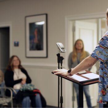 A patient sharing his story in front of an audience at a podium in an office setting with inspirational pictures on the walls